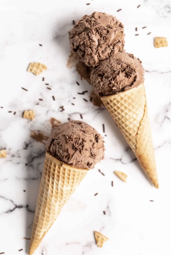 Two ice cream cones on a marble surface with chocolate ice cream and chocolate sprinkles.